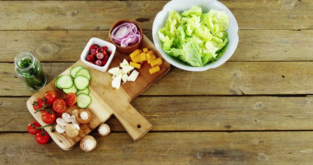 Table with fresh ingredients for a mixed salad, including cherry tomatoes, cucumbers, lettuce, red onions, cheese chunks, and a small jar of herbs and vinegar. Perfect for advertising healthy eating habits, organic food blogs, cooking tutorials, or fresh produce catalogs.