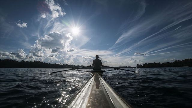 Sculler powering through sunlit lake with dramatic cloudscape and reflective water