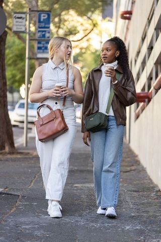 Two women walking and chatting on city sidewalk holding takeaway coffee cups, casual street style
