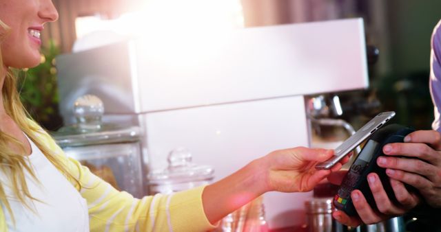 A young woman is making a contactless payment using her smartphone at a cafe. The scene depicts a modern and convenient transaction process. This is perfect for marketing materials related to digital payments, technology in retail, and modern consumer habits.