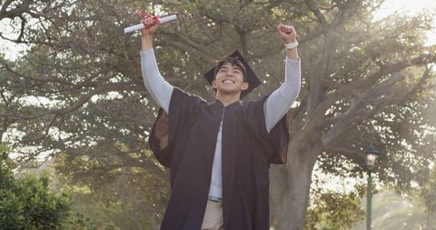 Asian graduate celebrating with diploma raising fists on sunlit campus lawn wearing cap and gown
