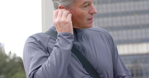 Senior man adjusting earbud while commuting on urban sidewalk with city buildings