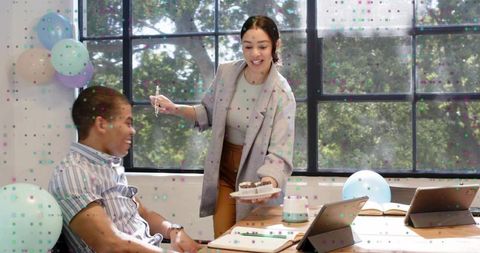 Office celebration with woman offering cake to colleague, balloons, confetti and tablets