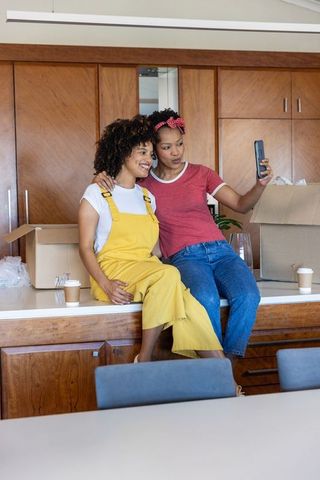 Diverse women relaxing on kitchen counter taking selfie while unpacking boxes in new home