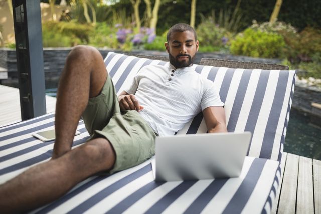 Man enjoying leisure time on a striped lounge chair while using a laptop outdoors. Ideal for illustrating remote work, modern lifestyle, outdoor relaxation, and casual summer activities. Suitable for blogs, articles, and advertisements related to technology, work-life balance, and outdoor living.