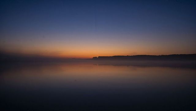 Twilight reflecting on calm lake with treeline silhouette and soft mist