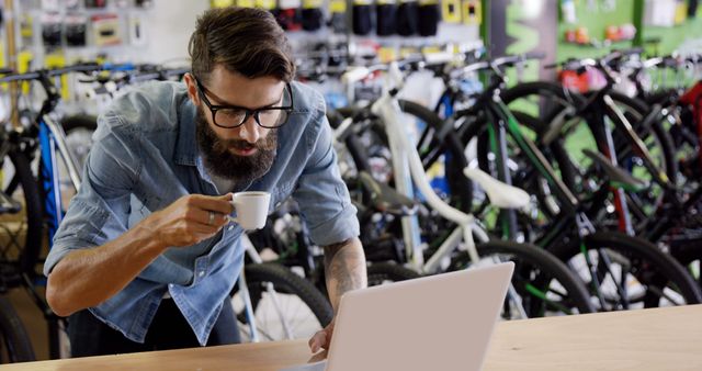 Man wearing glasses and a denim shirt drinks coffee while working on a laptop in a bicycle shop. Ideal for use in blogs about multitasking, managing retail business, planning and online work.
