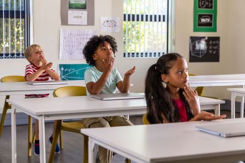 Diverse students raising hands and participating in bright classroom with laptops