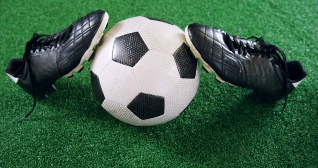 A classic black and white soccer ball is flanked by a pair of soccer cleats on a green artificial turf, with copy space. This setup evokes the excitement and passion associated with the sport of soccer.