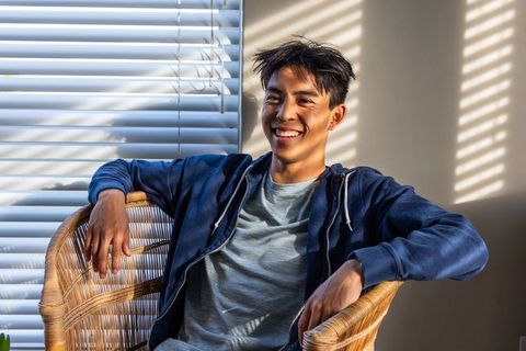 Asian man relaxing in rattan chair smiling under striped sunlight and blinds