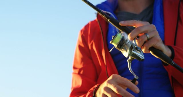 Man securing line on fishing rod wearing red jacket and standing outdoors. Image can be used for articles on outdoor hobbies, fishing tutorials, or advertisements for fishing gear.