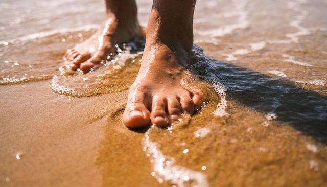 Standing barefoot on wet sand, foamy wave lapping over toes and soles