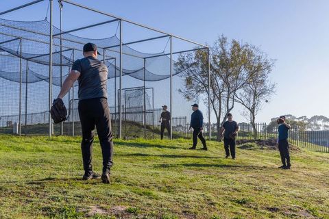 Men practicing baseball fielding drills on grassy slope beside batting cage in park
