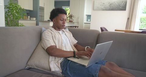 Young african american man working remotely on laptop while relaxing on modern cozy sofa