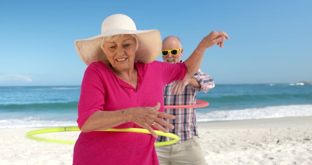 Two seniors having fun on the beach using hula hoops on a sunny day. Both are wearing casual clothing; the woman has on a pink shirt and a sunhat, while the man sports sunglasses and a plaid shirt. Ideal for illustrating concepts related to active aging, leisure, and healthful living. Perfect for use in articles, advertisements, and social media promoting senior fitness, beach vacations, and outdoor activities.