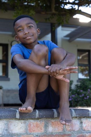 School-age african american boy sitting on brick wall on suburban porch barefoot thoughtful