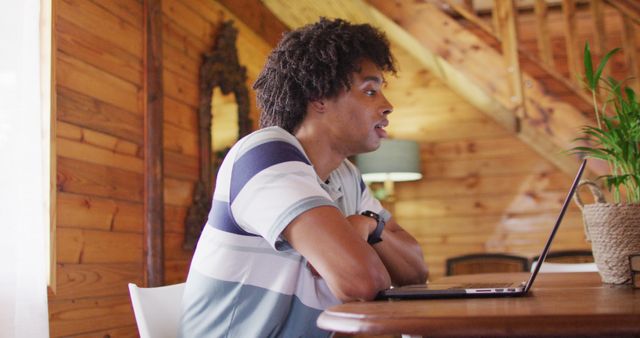 Young man sitting at wooden desk, working on laptop in home with wooden interior. Good for content about remote work, home office setups, freelancing, modern lifestyle, and technology in daily life.