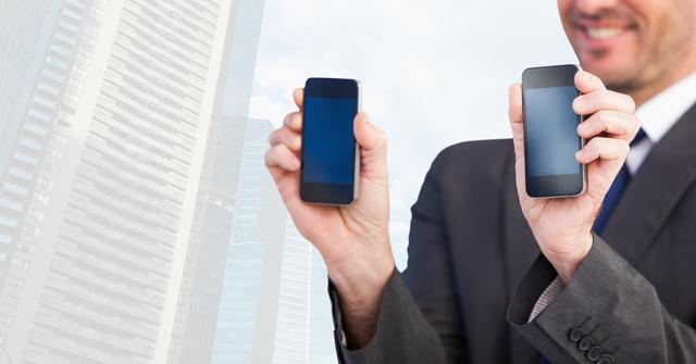 Midsection of smiling businessman holding smart phones with blank screens against skyscrapers. Useful for technology, business, and finance themes. Ideal for ads focusing on mobile technology, business solutions, or application development.