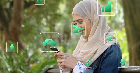 Young woman wearing hijab tapping smartphone summoning green eco-finance icons in park