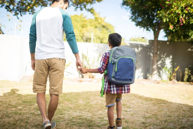 Rear view of father holding hand of son with backpack while walking in yard