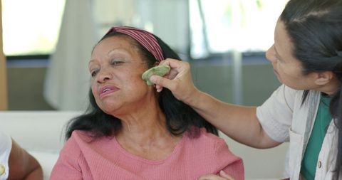 Senior african american woman receiving gentle jade gua sha facial massage from caregiver at home
