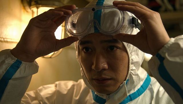 Asian lab worker adjusting goggles in white taped-seam coverall under warm lamp light