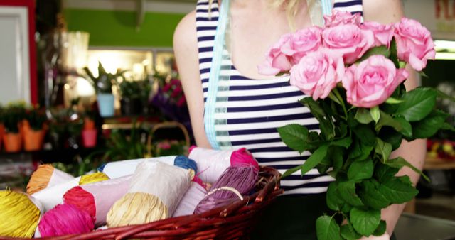 Woman holding bouquet of pink roses in one hand and basket of colorful wrapping papers in other hand, standing in flower shop. Ideal for use in articles about floristry, gift wrapping, floral shops, and florist tools.