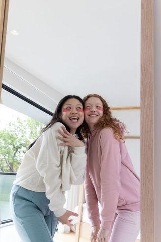 Asian and caucasian friends laughing in hoodies by mirror in minimal bright apartment