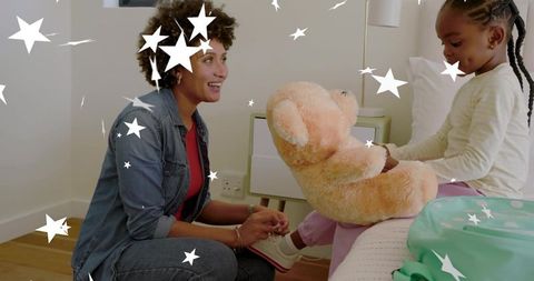 Caregiver kneeling fastening child's shoe while child holds teddy bear on bedroom bed