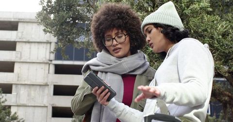 Young women checking smartphone while sharing electric scooter near urban parking garage