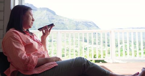 Middle-aged asian woman relaxing on balcony holding smartphone, mountain view