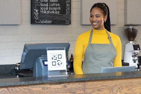 Smiling african american barista at counter with qr code pos and espresso machine