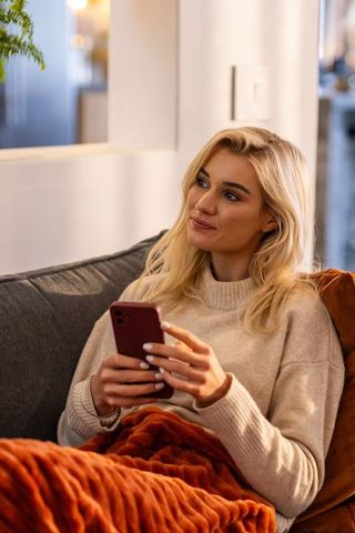 Young woman relaxing on sofa using smartphone under cozy blanket in modern living room