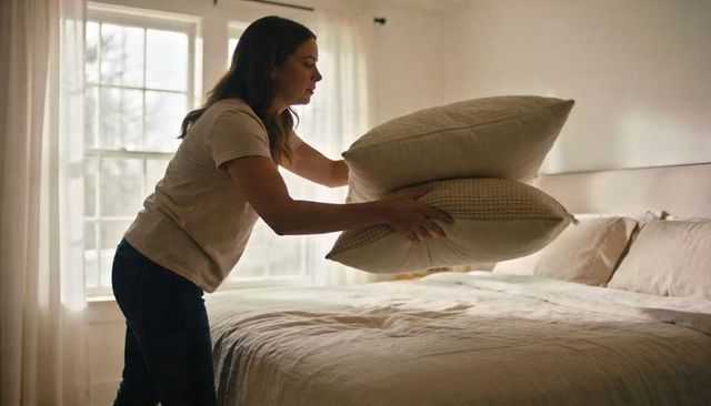 Woman placing decorative pillows on sunlit neutral bed for cozy morning