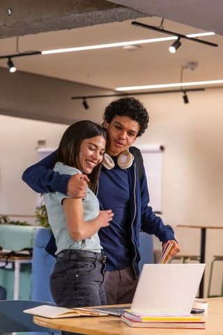 Teenage couple studying and smiling in campus lounge, collaborating on laptop and notebooks