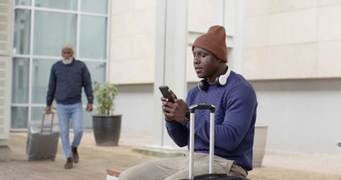 Young African American traveler checking phone while waiting with suitcase and headphones