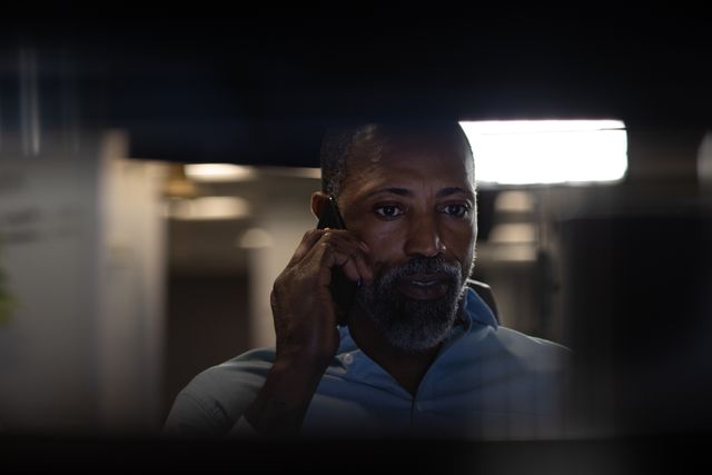 African American businessman working late in a modern office, talking on a smartphone and looking through a window. Ideal for use in articles about business, corporate culture, dedication, technology in the workplace, and professional communication.