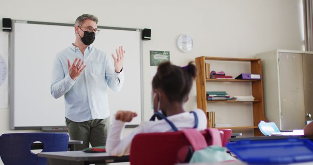 Male teacher wearing mask teaching diverse students in an elementary school classroom. Potential uses include educational materials, school advertising, articles about pandemic precautions in education, classroom management resources, and health and safety guidelines in schools.