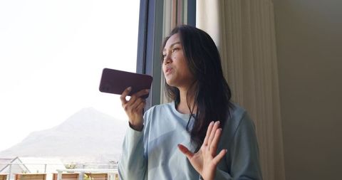 Asian woman speaking into smartphone standing by balcony window with mountain view