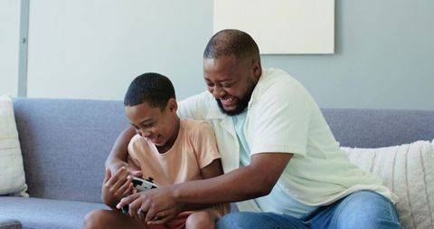 African american father and son laughing and playing video game on sofa bonding moment