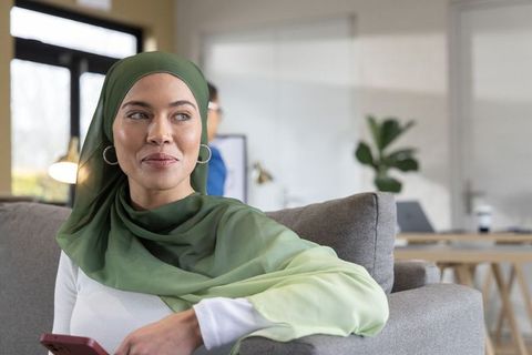 Confident woman in green hijab holding smartphone and smiling in contemporary coworking lounge