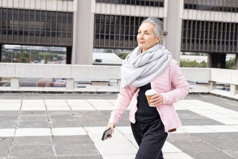 Mature woman walking on urban terrace wearing scarf holding coffee cup and smartphone