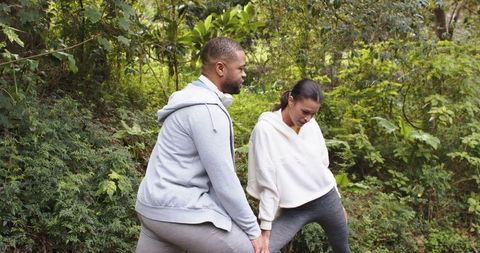 Diverse couple stretching on forest trail in activewear with headphones, outdoor fitness