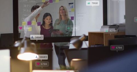 Diverse team planning on glass wall with sticky notes, tablet and social reactions