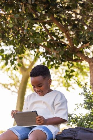 African american boy using tablet under sunlit tree outdoors smiling, learning, relaxing