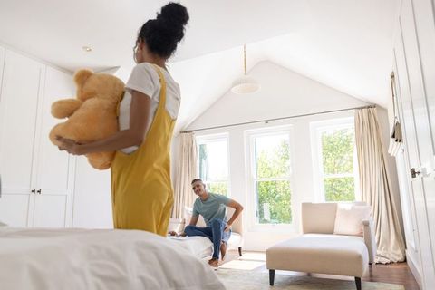 Couple unpacking teddy bear in sunlit vaulted bedroom, playful intimate home moment