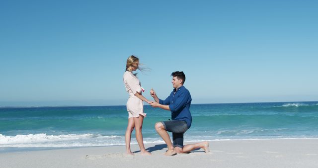 A man is kneeling on the sandy beach presenting an engagement ring to his partner against a backdrop of a clear blue ocean and sky. The couple displays emotions of love and excitement. Perfect for themes related to romance, engagements, proposals, beach weddings, love stories, and surprise moments.