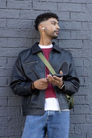 Indian man standing by gray brick wall holding smartphone wearing earbuds leather jacket