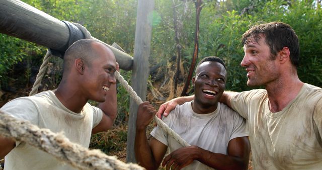 Three young men, one Asian, one African American, and one Caucasian, are laughing and enjoying a moment of camaraderie outdoors. Their joyful expressions and dirty clothes suggest they've been engaged in some kind of physical, team-building activity.