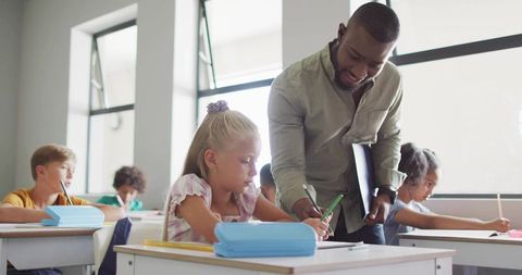 Male teacher guiding elementary student writing at desk in bright classroom with windows